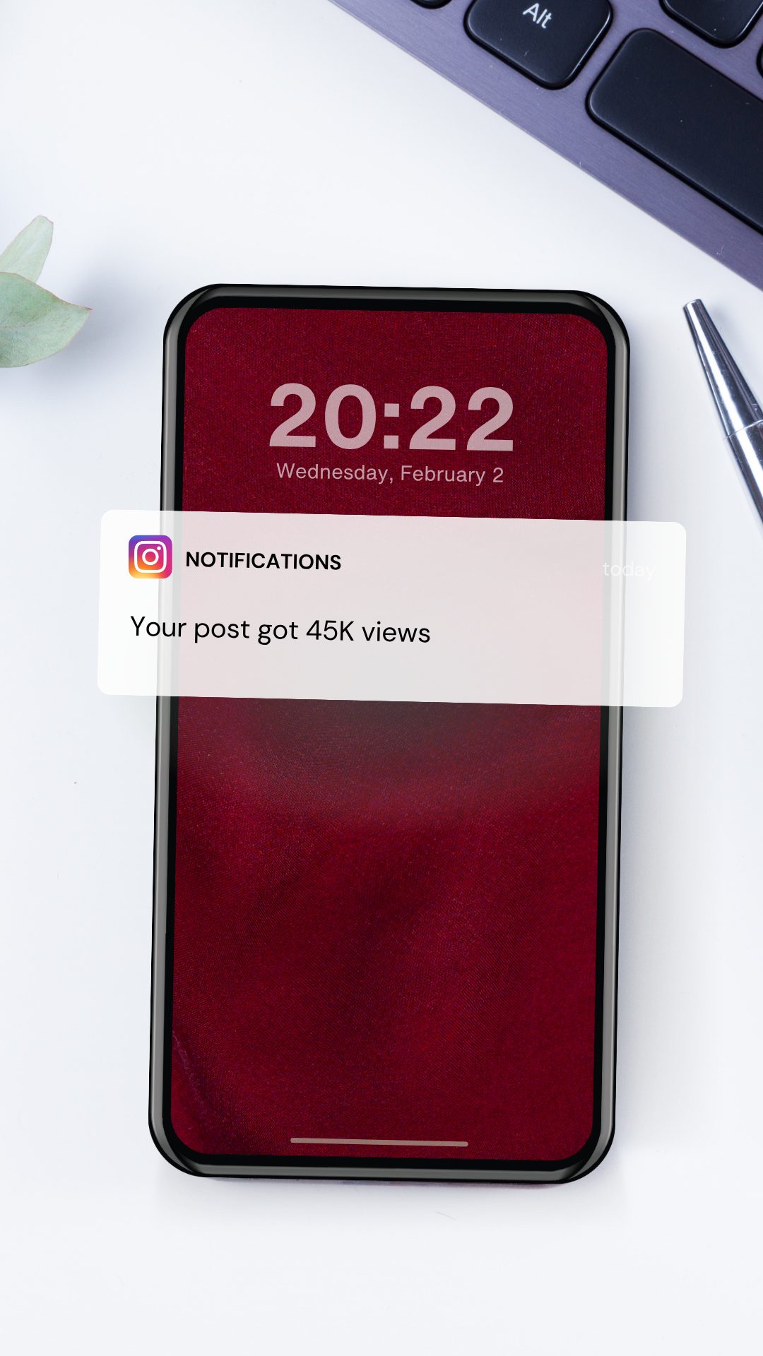 Smartphone on a white desk showing a burgundy lock screen with an Instagram notification reading &ldquo;Your post got 45K views&rdquo;